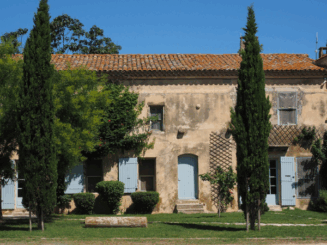 Casa rural en finca rústica con fachada de piedra, contraventanas azules y jardín con cipreses.